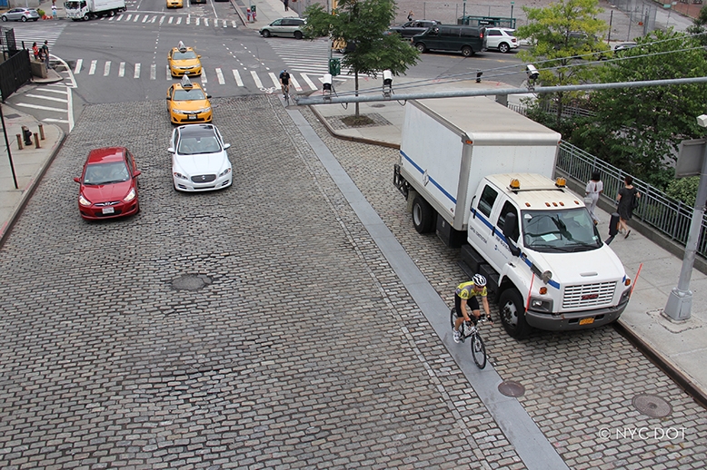 Cyclist and drivers traversing down street made of cobblestone